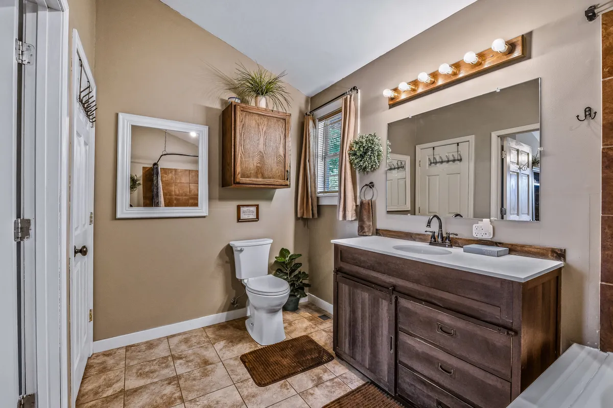 A tranquil bathroom with a vanity and sink, large mirror, toilet, and a window with curtains. The room is inviting and bright, suggesting comfort and practical convenience.
