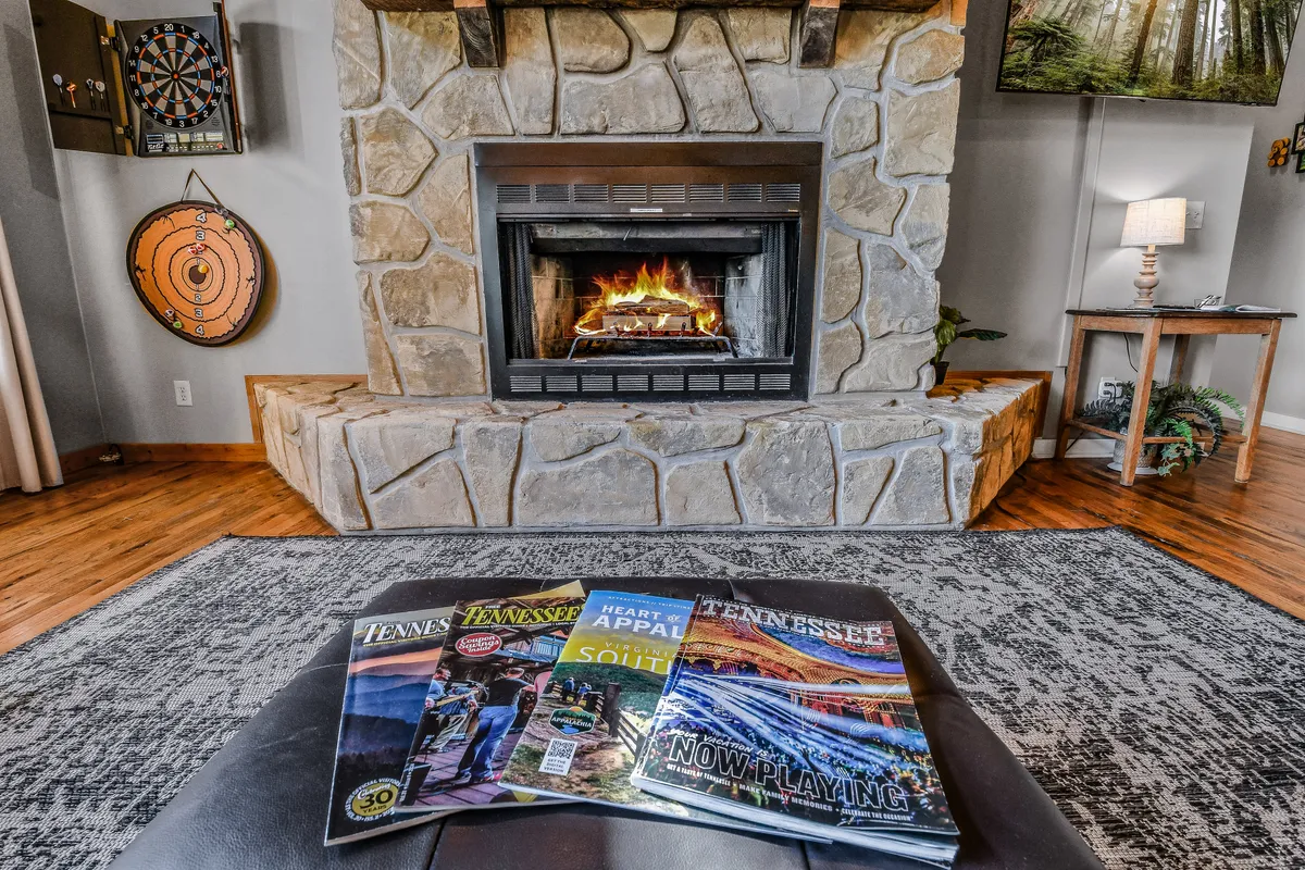 Inside the cabin, this image highlights a cozy living area with a crackling fireplace, a stone mantel, a dartboard, and a coffee table with magazines. The space encourages indoor social activities like relaxing by the fire, playing games, or enjoying friendly conversation.