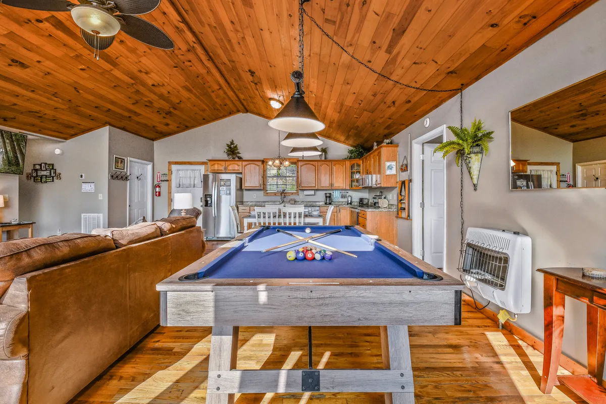 This image features a spacious game room with a regulation pool table as its centerpiece. Comfortable lounge seating is arranged nearby, and the room is anchored by warm wooden ceilings and an open kitchen area in the background. The space exudes a casual, inviting atmosphere perfect for group entertainment and relaxation.