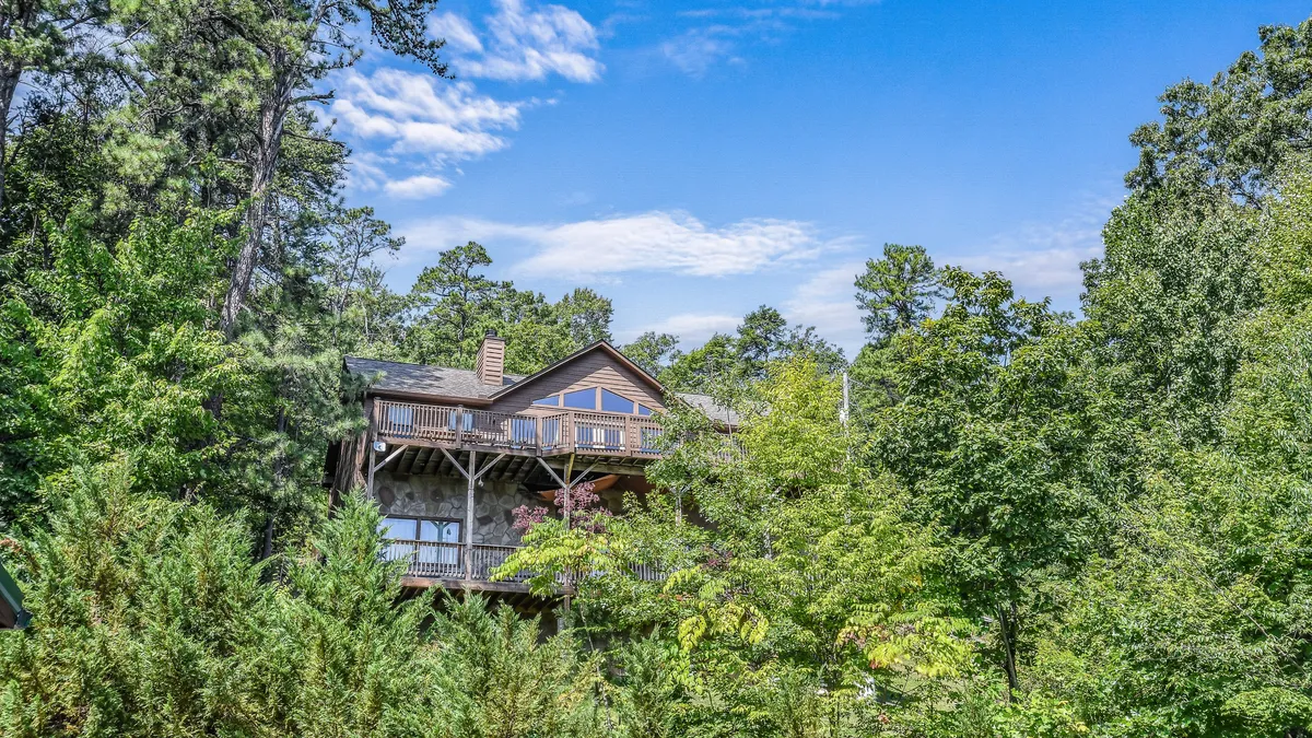 This exterior image features the cabin nestled among tall trees, with a balcony visible and a peaceful natural setting surrounding the home. The scene highlights an inviting and accessible outdoor area suitable for all ages and mobility levels, with ample space for gathering and relaxation.