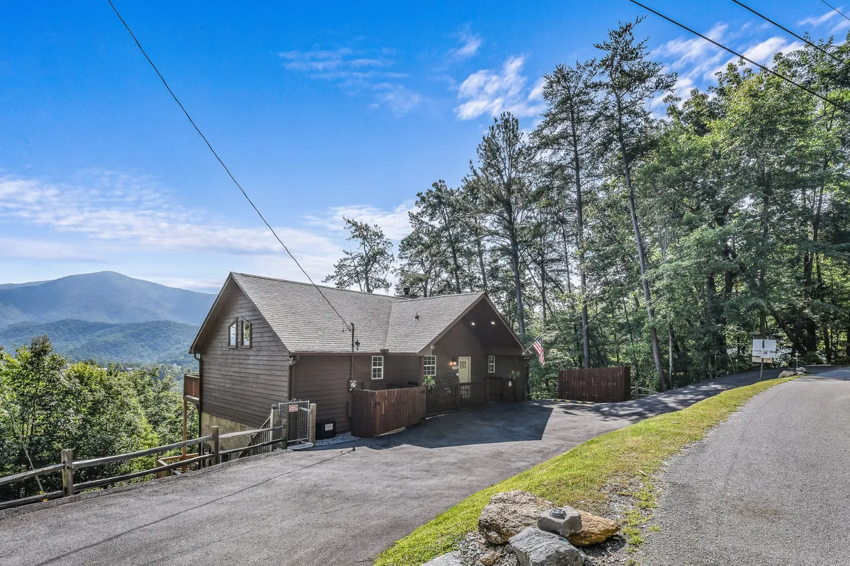 This image features the exterior of the cabin set against a stunning mountain backdrop, framed by tall trees and a spacious driveway. The tranquil setting hints at the natural adventures awaiting families, including nature walks and scenic viewing.