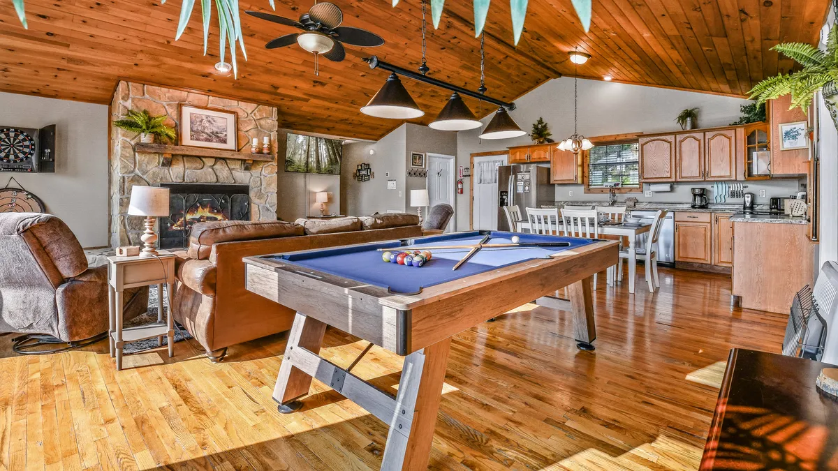 An inviting dining area adjacent to a kitchen, featuring a pool table for entertainment, a cozy fireplace, and a wooden ceiling. The dining table sits ready for a lively family meal amidst a relaxed, social setting.