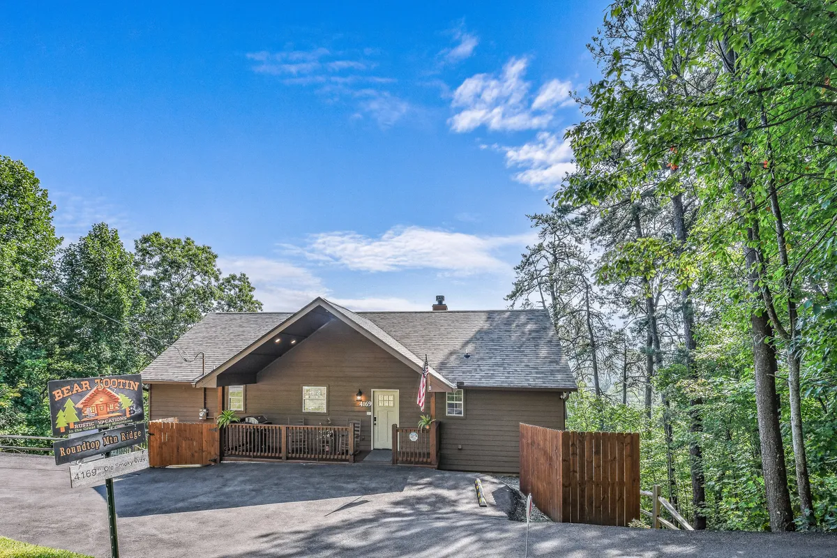 The image highlights the warmly lit wooden facade of the house, a spacious porch, and a welcoming sign, all set amidst tranquil natural surroundings. The scene communicates openness and an inviting entryway for new arrivals.