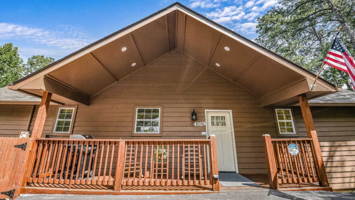 This image features the front porch of the cabin, with welcoming furniture, an American flag, and a wooden railing. The porch offers a scenic spot for relaxing, mingling with friends, or simply soaking in the ambiance of the surroundings.