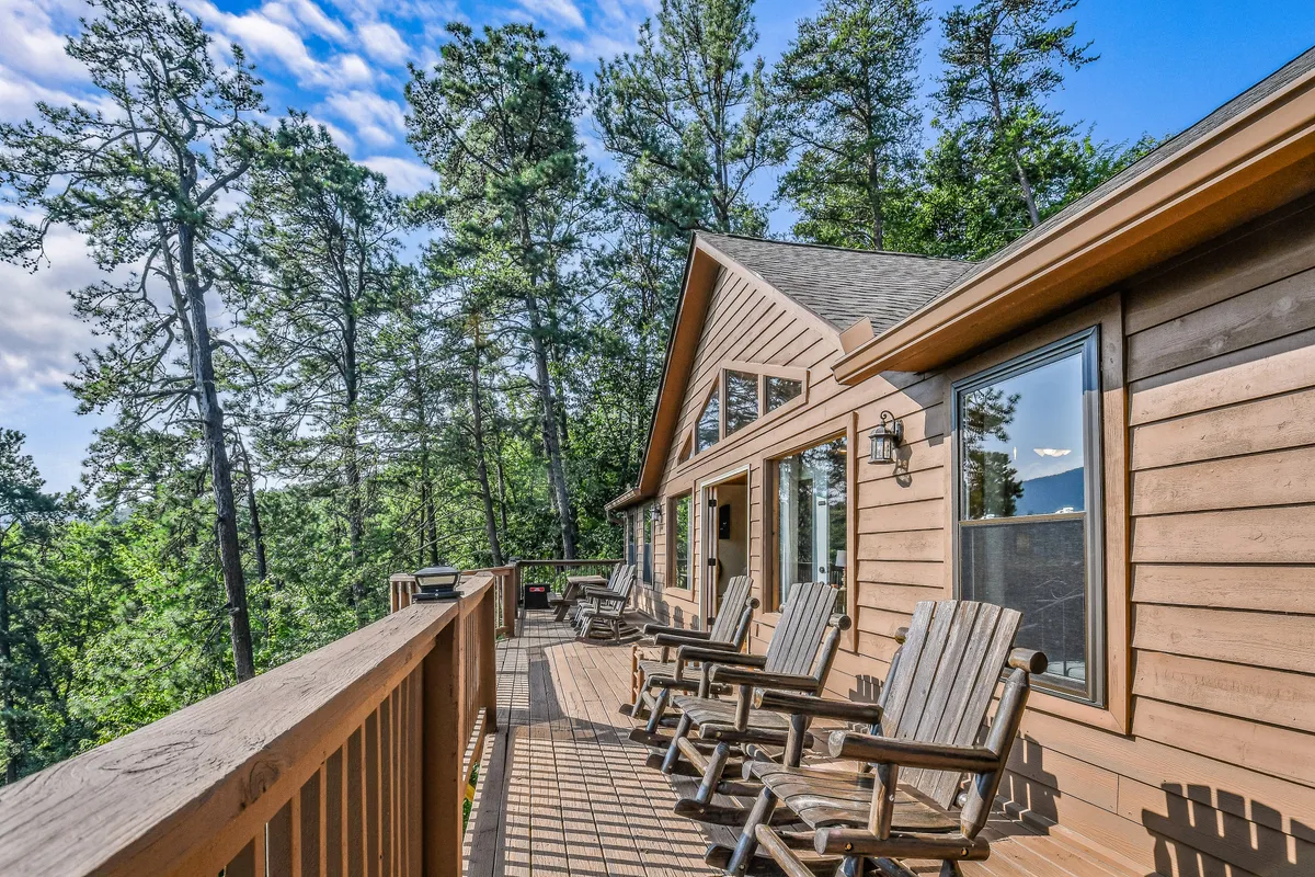 This image shows a spacious wooden deck with reclining chairs, surrounded by lush trees and large windows that connect the indoor living spaces with the outdoors. The setting is ideal for relaxing, socializing, or hosting group gatherings while enjoying the peaceful mountain atmosphere.