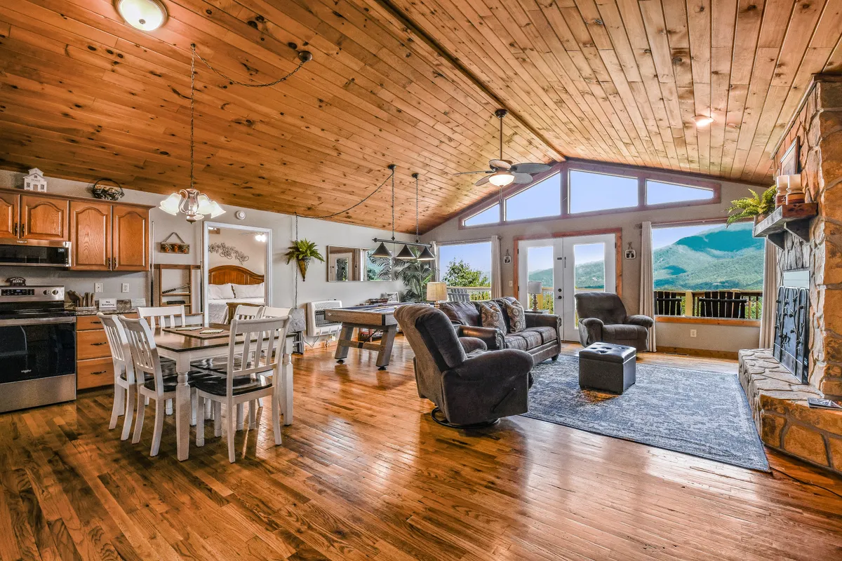 A spacious, inviting living room with large windows framing sweeping mountain vistas. The room features a wooden ceiling, a handsome stone fireplace, a dining table with chairs, and a comfortable seating area. Sunlight streams in, highlighting the connection to the outdoors and the warmth of the space.