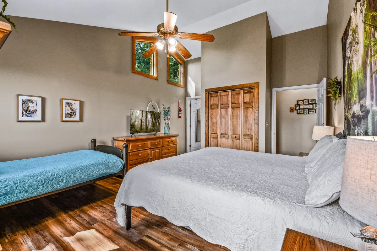 A well-lit bedroom with a queen-sized bed, a twin bed, wooden dresser, decorative art frames, and a ceiling fan. The room exudes a welcoming and cozy atmosphere, ideal for restful family stays.