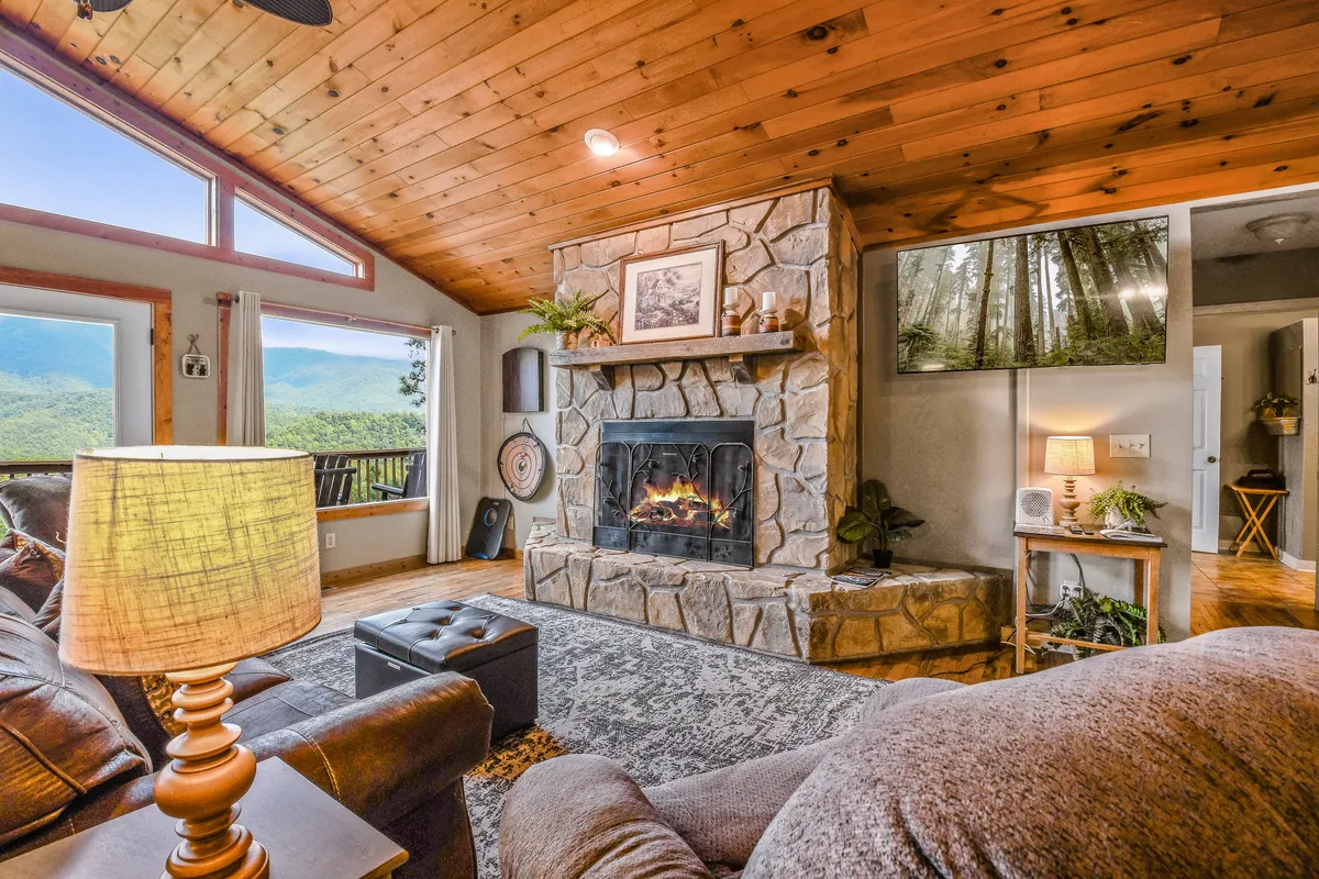 A comfortable living room setup with a plush seating arrangement facing a stone fireplace, large windows that overlook the mountains, and a flat-screen TV displaying a serene nature image. The room radiates warmth and comfort, ideal for movie nights or relaxing by the fire.