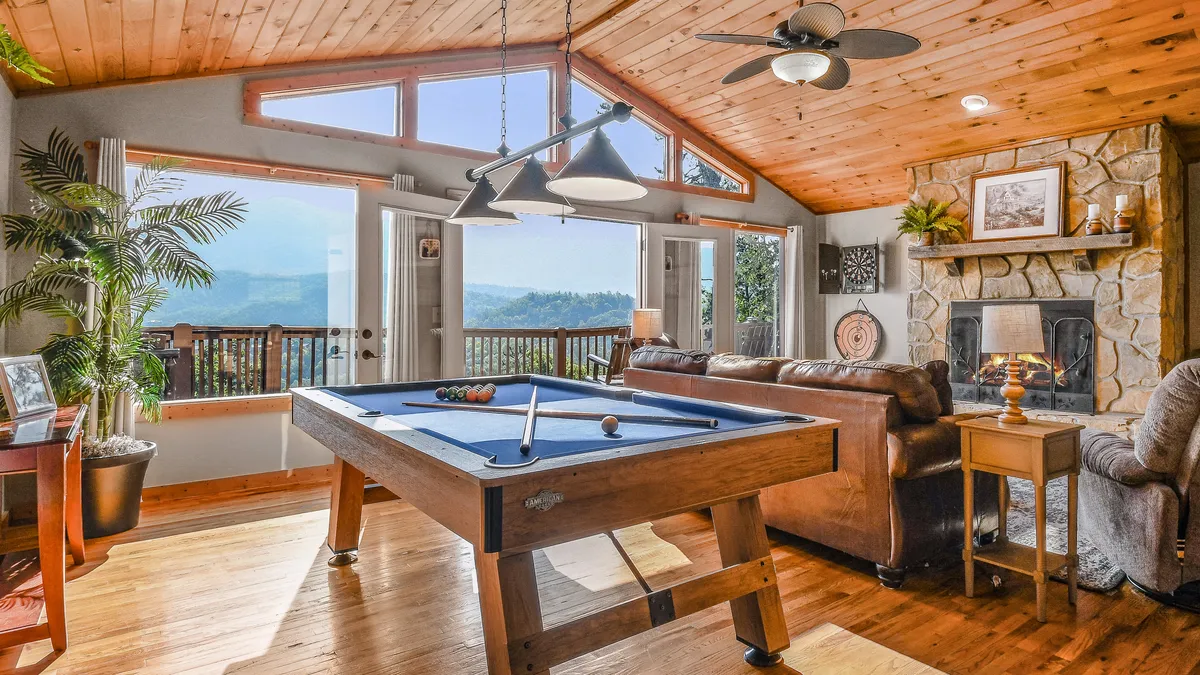 The image showcases the inviting interior of a mountain cabin, where a pool table takes center stage beneath soaring ceilings, flanked by expansive windows framing stunning mountain vistas. A rustic stone fireplace adds cozy warmth, while natural light bathes the space, creating a welcoming atmosphere perfect for gatherings and friendly games.