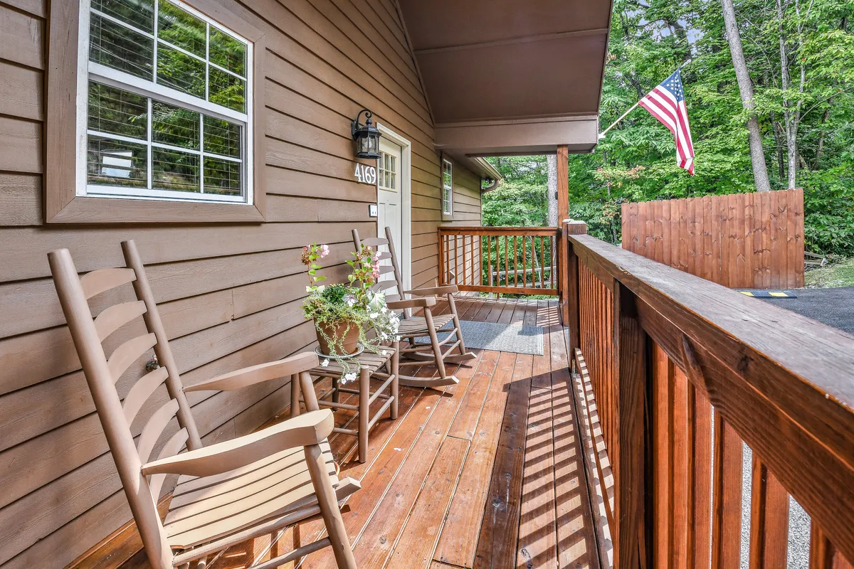 This image displays a classic wooden porch with rocking chairs, potted plants, a flag, and sturdy railing, set against a backdrop of inviting greenery. The porch exudes a sense of welcome and relaxation, offering a glimpse into the property's charming outdoor living space.
