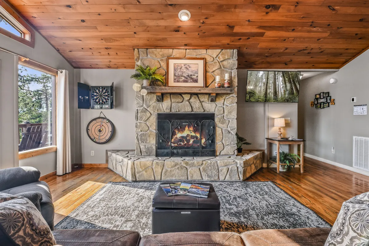 A cozy room showcasing a stone fireplace, a dartboard mounted on the wall, and large windows letting in natural light. A coffee table and seating area invite guests to relax, play games, or read, accentuating the space's versatility for all ages.