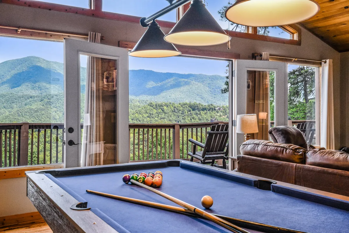 This inviting interior captures a game area with a pool table positioned near large windows that offer a stunning mountain view. A plush sofa and a dining area provide ample space for relaxing, playing games, or sharing stories, enhancing the sense of togetherness and entertainment for all ages.