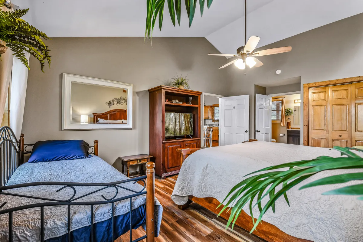 This image displays two beds—one with blue bedding and one with beige bedding—set in a cozy bedroom with wooden furniture, a large mirror, and a tall potted plant. The room is softly lit and feels spacious, emphasizing relaxation and comfort.
