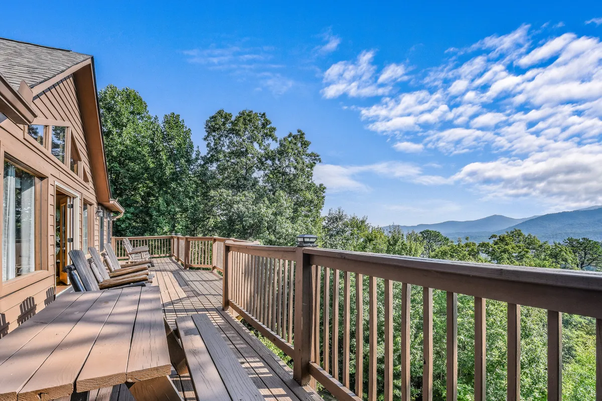 A wooden deck extends toward a stunning panoramic view of the mountains, framed by surrounding trees. The peaceful mood invites guests to relax outdoors, watch the sunrise, or stargaze in the mountain air.